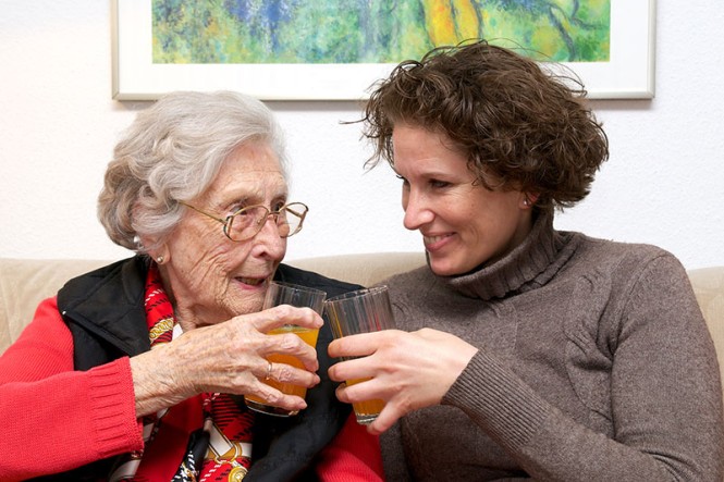 Eine ältere Dame und eine pflegende Angehörige stoßen mit zwei Wassergläsern an