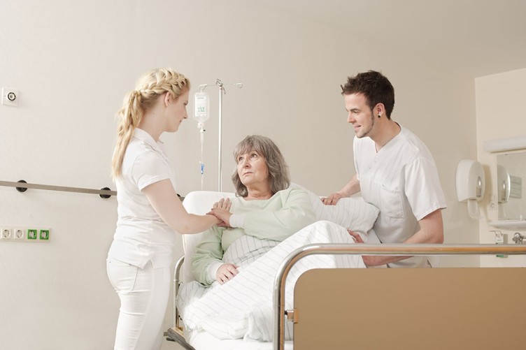 Eine Pflegerin und ein Pfleger stehen am Krankenhausbett einer älteren Dame