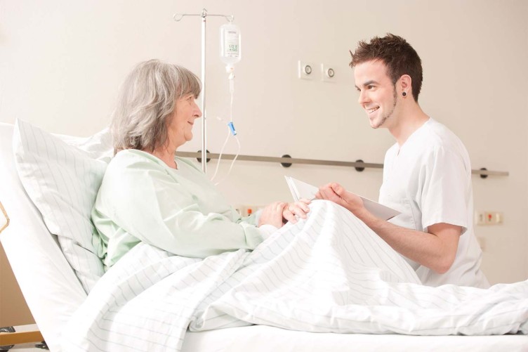 Ein Pfleger spricht mit einer älteren Frau, die im Krankenhausbett liegt.