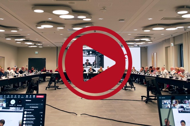 Ein Sitzungsraum mit vielen Teilnehmenden in einer Diskussion, einige davon sind virtuell zugeschaltet. Darüber ist ein großer roter Button mit einem Play-Symbol.
