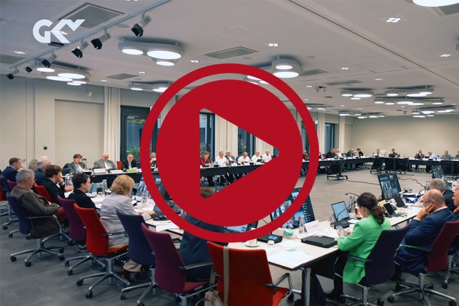 Viele Menschen sitzen an einem großen O-förmigen Tisch in einem Konferenzraum. Auf dem Bild ist davor ein großer roter Play-Button.
