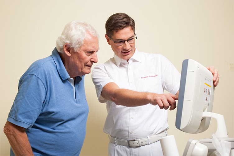 Ein Arzt und ein älterer Mann schauen zusammen auf einen Untersuchungsbildschirm. Der Arzt zeigt etwas.