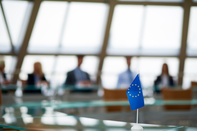 Eine kleine Europa-Flagge auf einem Tisch. Im Hintergrund sitzen Menschen an Konferenztischen