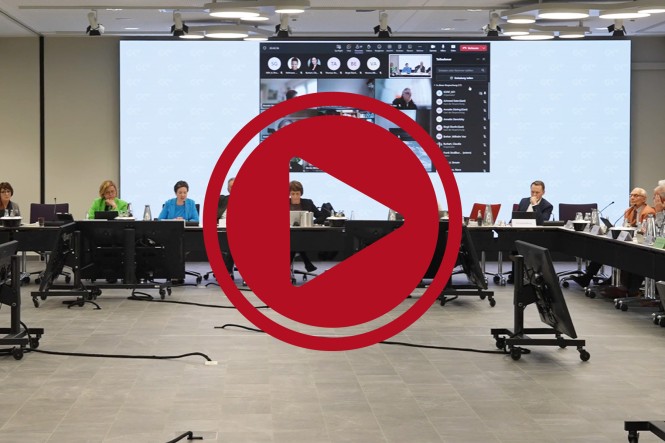Das Präsidium der Mitgliederversammlung. Auf dem Bild ist davor ein großer roter Play-Button.