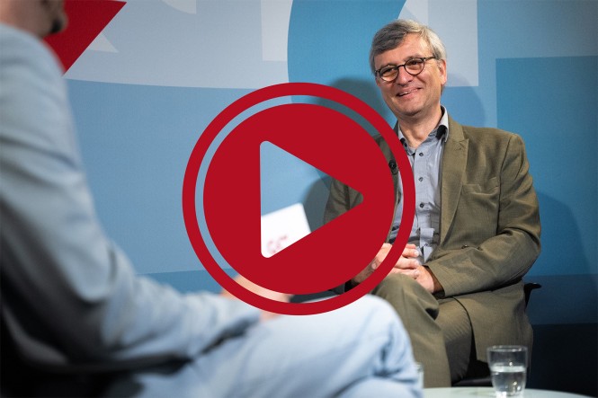 Der Interviewgast, Dr. Ramin Tavakolian, sitzt in einem Stuhl. Ein großer roter Play-Button ist davor zu sehen.