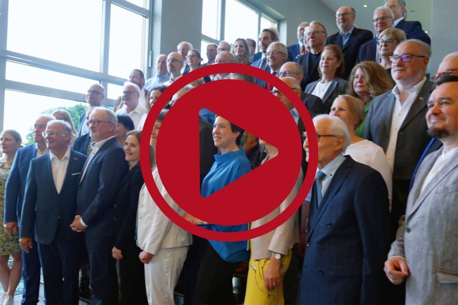 Ungefähr 50 Menschen stehen an einer Treppe zu einem Gruppenfoto zusammen. Davor ist ein großer roter Play-Button eingefügt.
