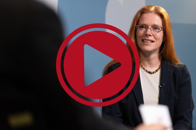 Friederike Kuhnt, Referatsleiterin beim GKV-Spitzenverband, im Gespräch mit einer anderen Person. Davor ist ein roter Play-Button eingefügt.