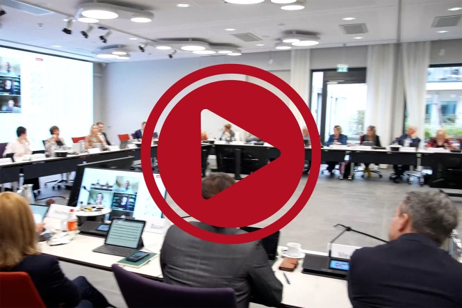 Mehrere Menschen sitzen in einem großen Besprechungsraum vor einer Leinwand. Davor ist ein roter Play-Button eingefügt.
