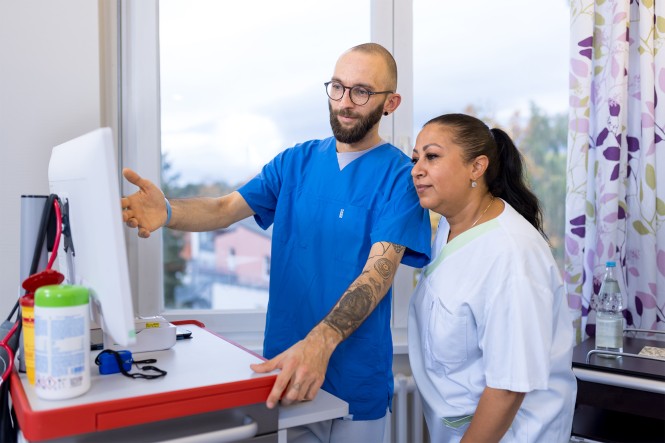 Ein männlicher Pfleger zeigt einer anderen Pflegekraft etwas auf einem Monitor. Die beiden stehen in einem Besprechungsraum im Krankenhaus