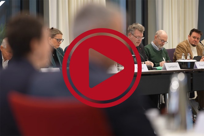 Mehrere Menschen sitzen in einem Sitzungsraum während einer Tagung. Davor ist ein roter Play Button zu sehen.