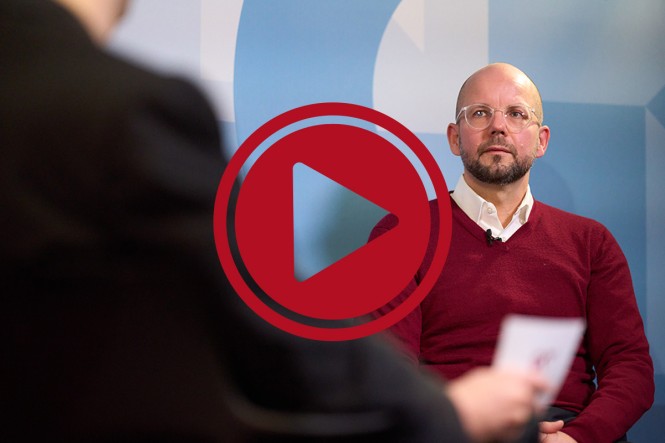 Dr. Ronny Klein, Fachreferent im GKV-Spitzenverband im Gespräch mit einer anderen Person. Davor ist ein roter Playbutton zu sehen.