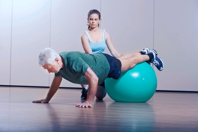 Eine Therapeutin trainiert mit einem älteren Mann auf einem Medizinball
