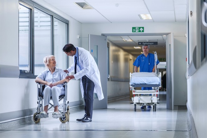 Ein Krankenhausflur. Ein Arzt spricht mit einer Patientin im Rollstuhl, ein Pfleger schiebt ein Krankenhausbett.