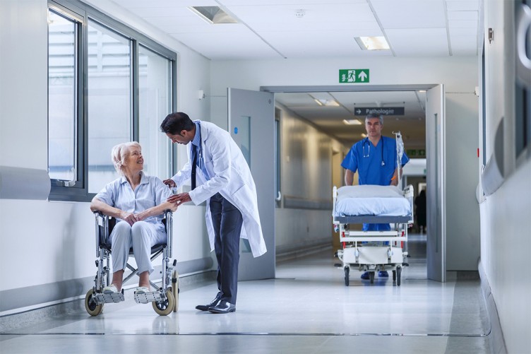 Ein Krankenhausflur. Ein Arzt spricht mit einer Patientin im Rollstuhl, ein Pfleger schiebt ein Krankenhausbett.