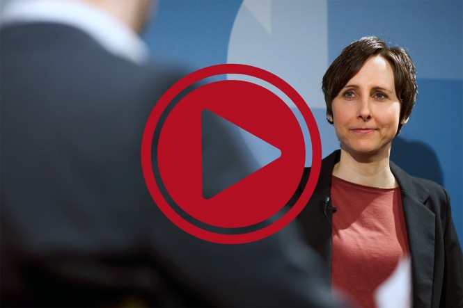 Eine Frau ist im Gespräch mit einer anderen Person, die man nur angeschnitten sieht. Im Vordergrund ist ein großes, rotes Playsymbol zu sehen.